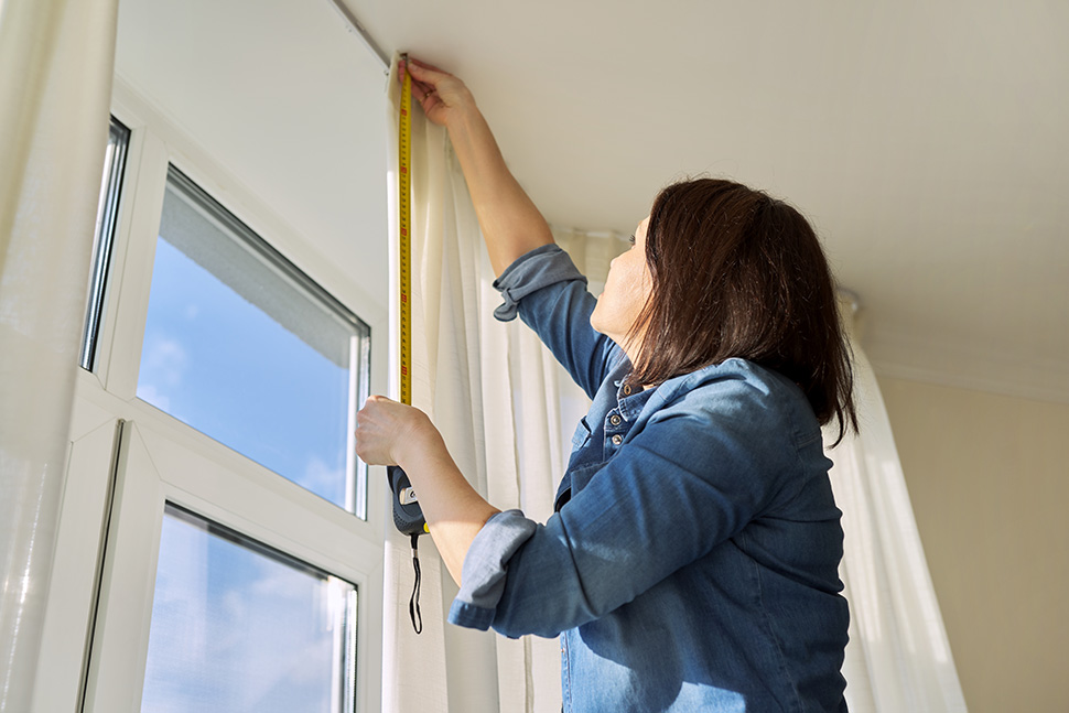Eine Frau in einem Jeanshemd misst mit einem Maßband ein Fenster in einem Raum.